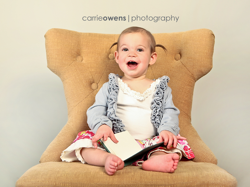sandy utah photographer captures smiling one year old girl reading a book on a chair